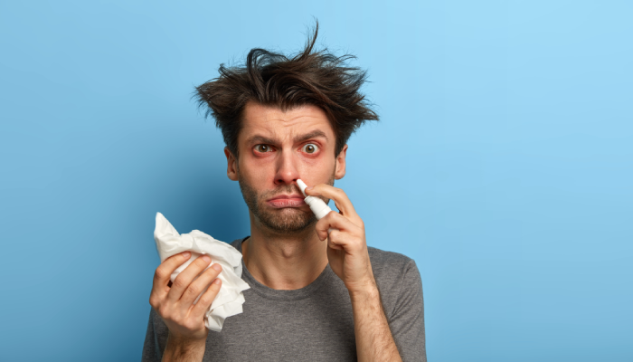 Fotografía para artículo del blog topfarma sobre antihistamínicos. Un hombre esteriliza su nariz tapada con un bote de spray y con la otra mano sostiene un pañuelo.